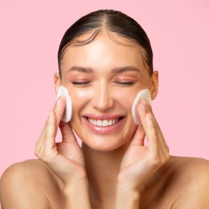 daily skincare. excited young lady cleaning face with cotton pads and smiling, doing skin care routine over pink background